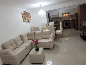 A modern living room with a beige sectional sofa, a glass coffee table, and a dining area with chairs.