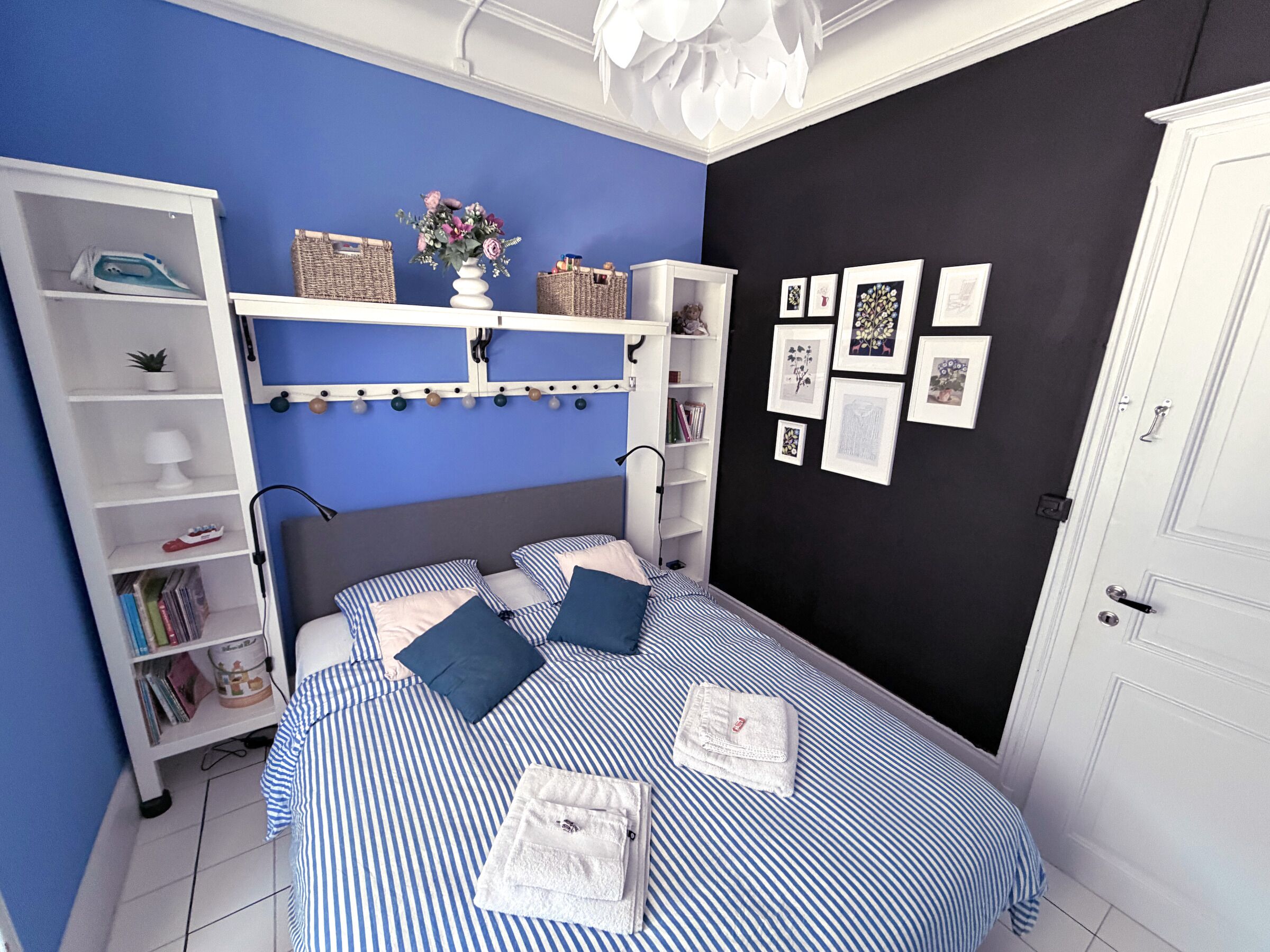 Une chambre à coucher comprenant un lit, une armoire, une étagère avec des livres et des objets décoratifs, et un mur orné de tableaux encadrés.