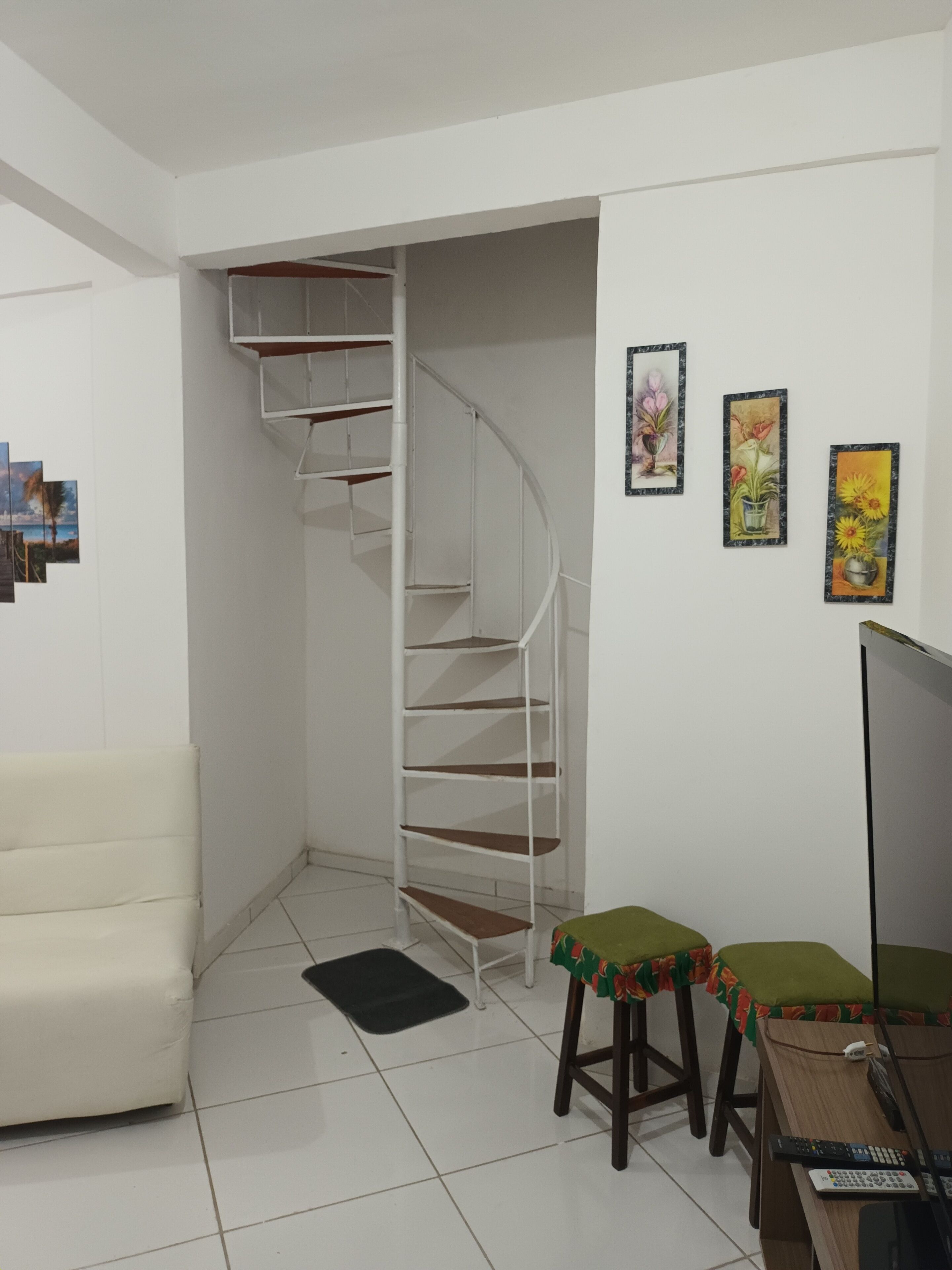 A room with a white spiral staircase, a white sofa, two stools with green cushions, and a desk with a computer and remote controls.