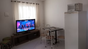 En stue med et fladskærms-tv, et spisebord med stole, et køleskab og et vindue med gardiner.