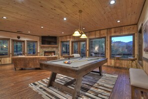 Un interior de madera con mesa de billar, chimenea de piedra y grandes ventanales que ofrecen vistas al exterior.