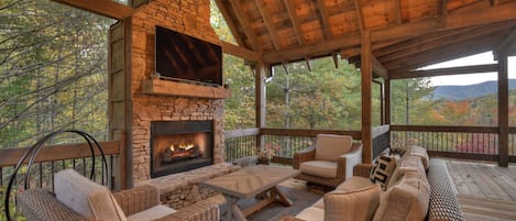 Un espacio de estar exterior techado con chimenea de piedra, muebles de mimbre y un televisor montado en la pared.