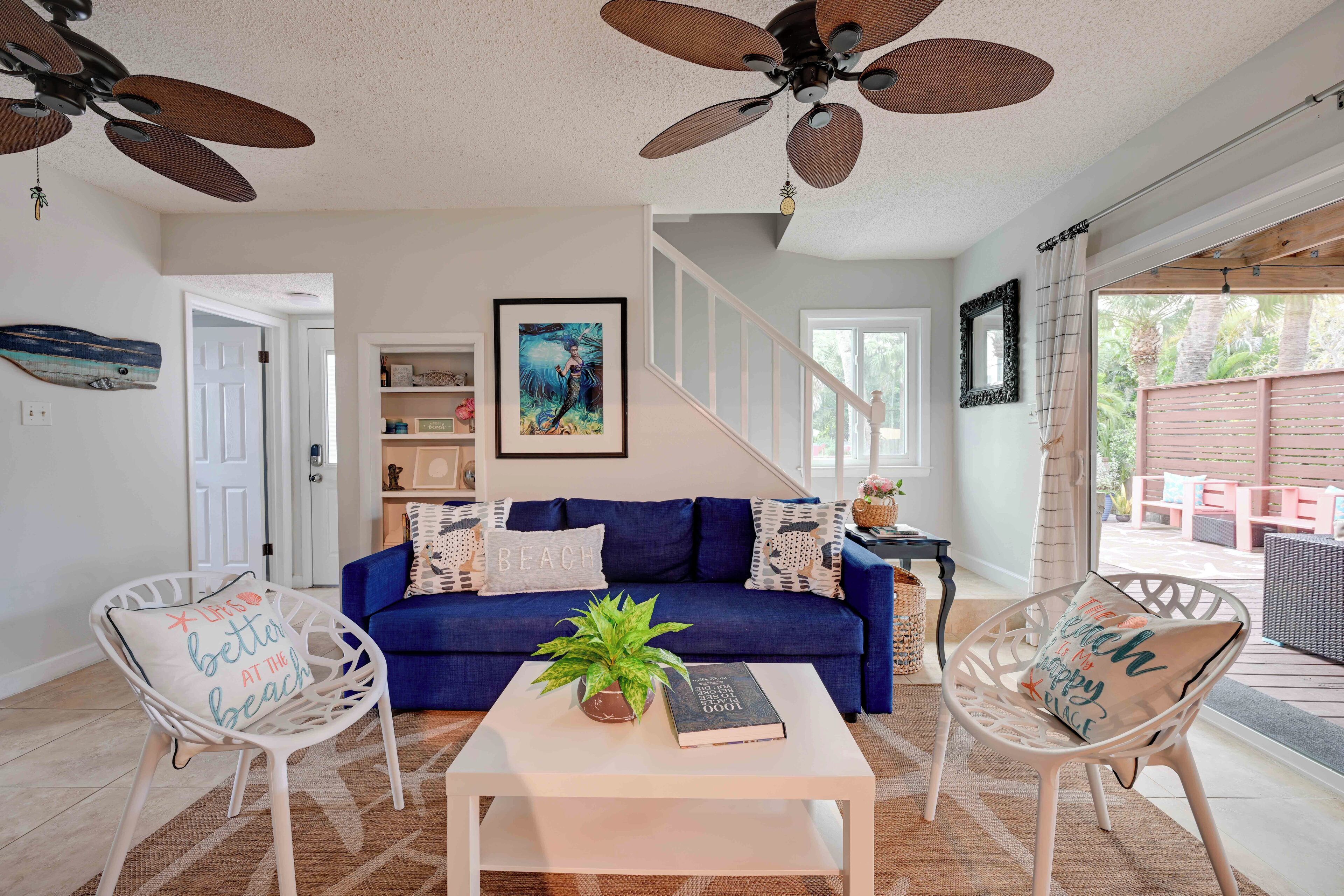 A living room with a blue sofa, white chairs, a coffee table with a plant and book, and ceiling fans.