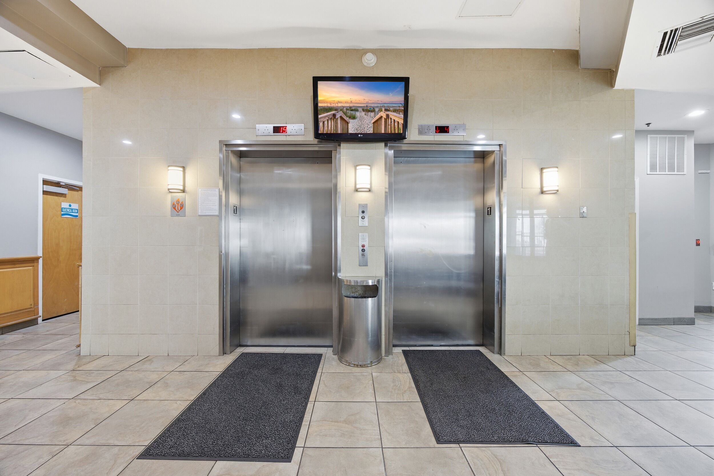 Een hotel lobby met twee liften, een digitale klok, een waterdispenser en een televisie die een mooi uitzicht toont.