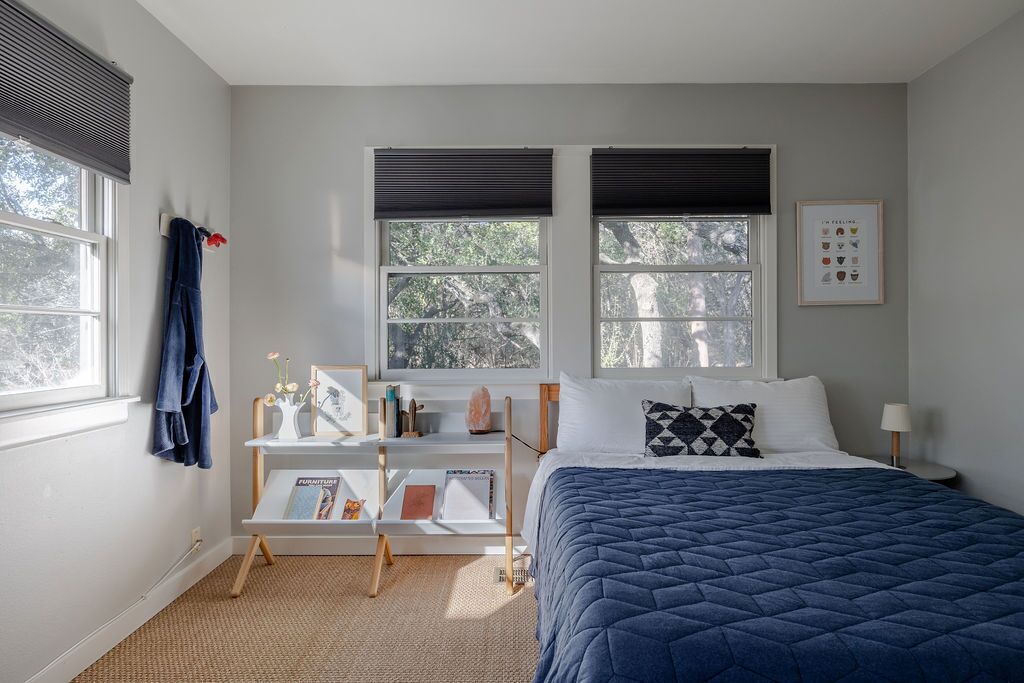 A bedroom with a bed, two windows with blinds, a bedside table with a lamp, a small shelf with books and decorative items, and a wall-mounted picture.