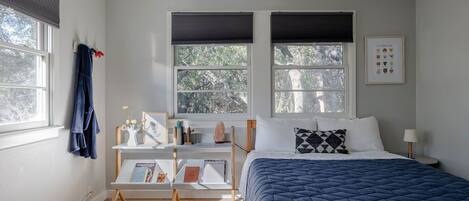 A bedroom with a bed, two windows with blinds, a bedside table with a lamp, a small shelf with books and decorative items, and a wall-mounted picture.