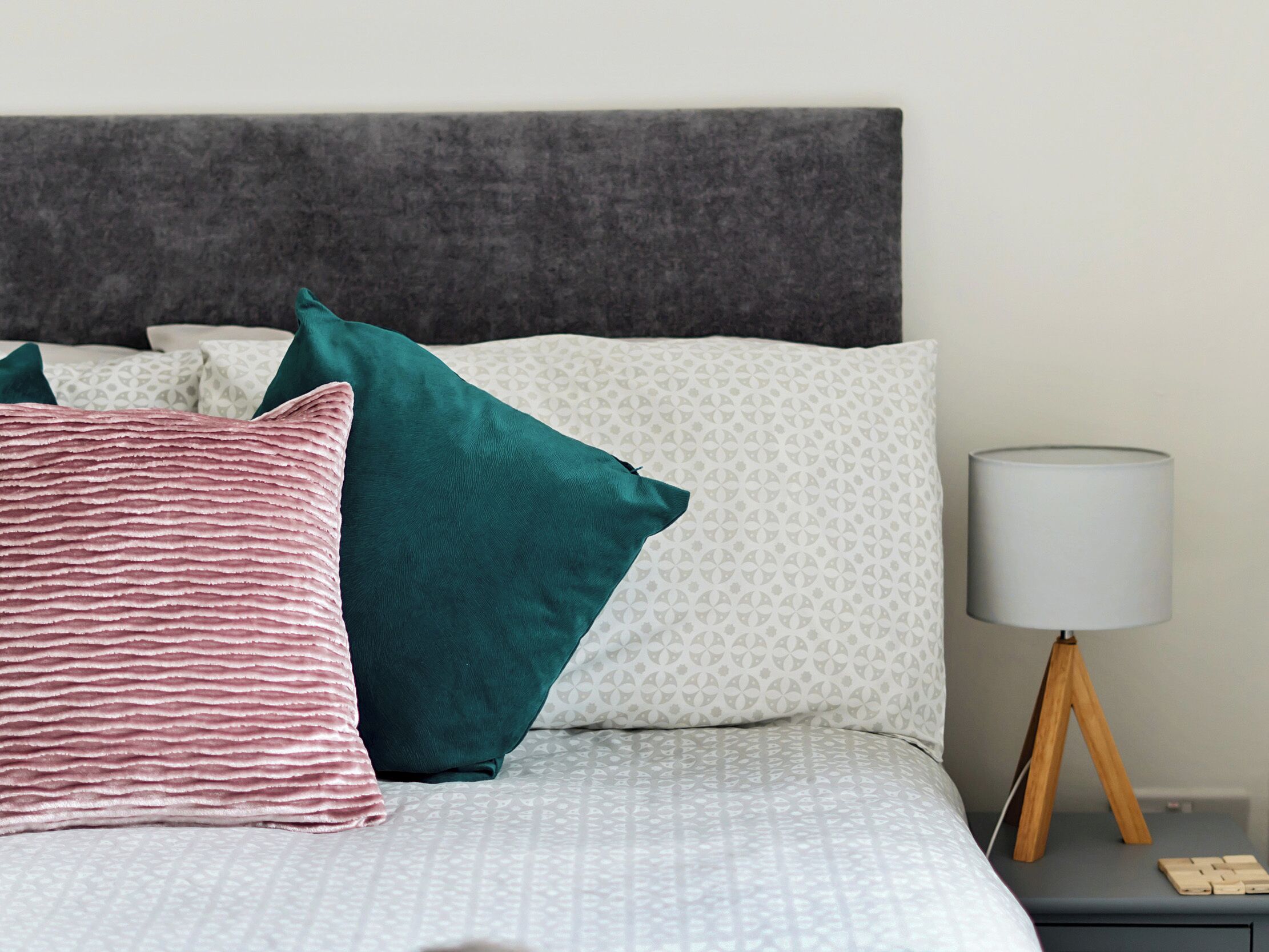 A neatly made bed with a patterned bedspread, multiple pillows in teal and white, and a bedside table with a lamp and a wooden stand.
