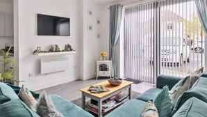 A modern living room with a flat-screen TV, a fireplace, a coffee table with books, and a view through large windows. - Mary Bowes Terrace (Bournemouth)