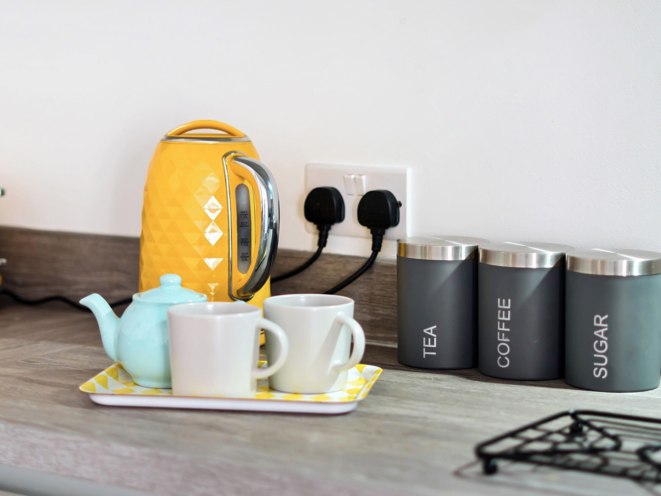 A kitchen counter with a yellow kettle, a blue teapot, and three containers labeled tea, coffee, and sugar.