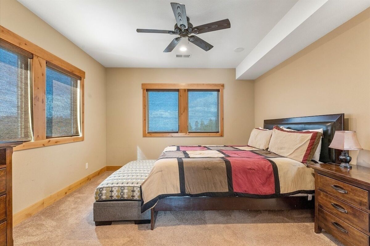 A bedroom with a bed, a ceiling fan, two windows, a nightstand, and a lamp.