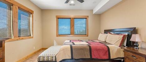 A bedroom with a bed, a ceiling fan, two windows, a nightstand, and a lamp.