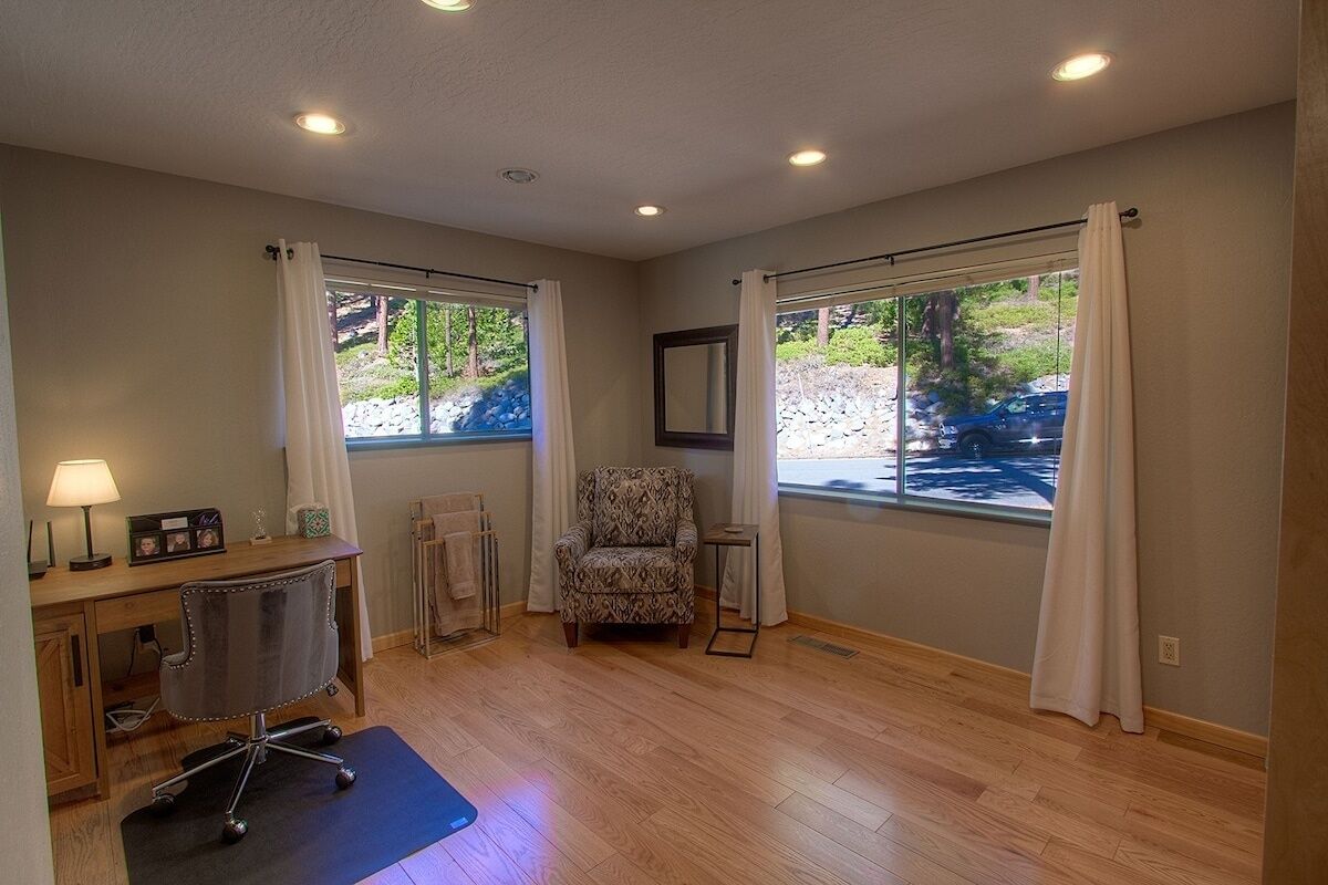 A room with a wooden floor, a desk with a chair, a small table, and a chair. There are two windows with curtains, a mirror on the wall, and a view of greenery outside.