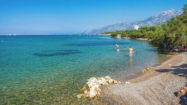 A clear coastal view with a pebble beach, calm sea, and distant mountains.