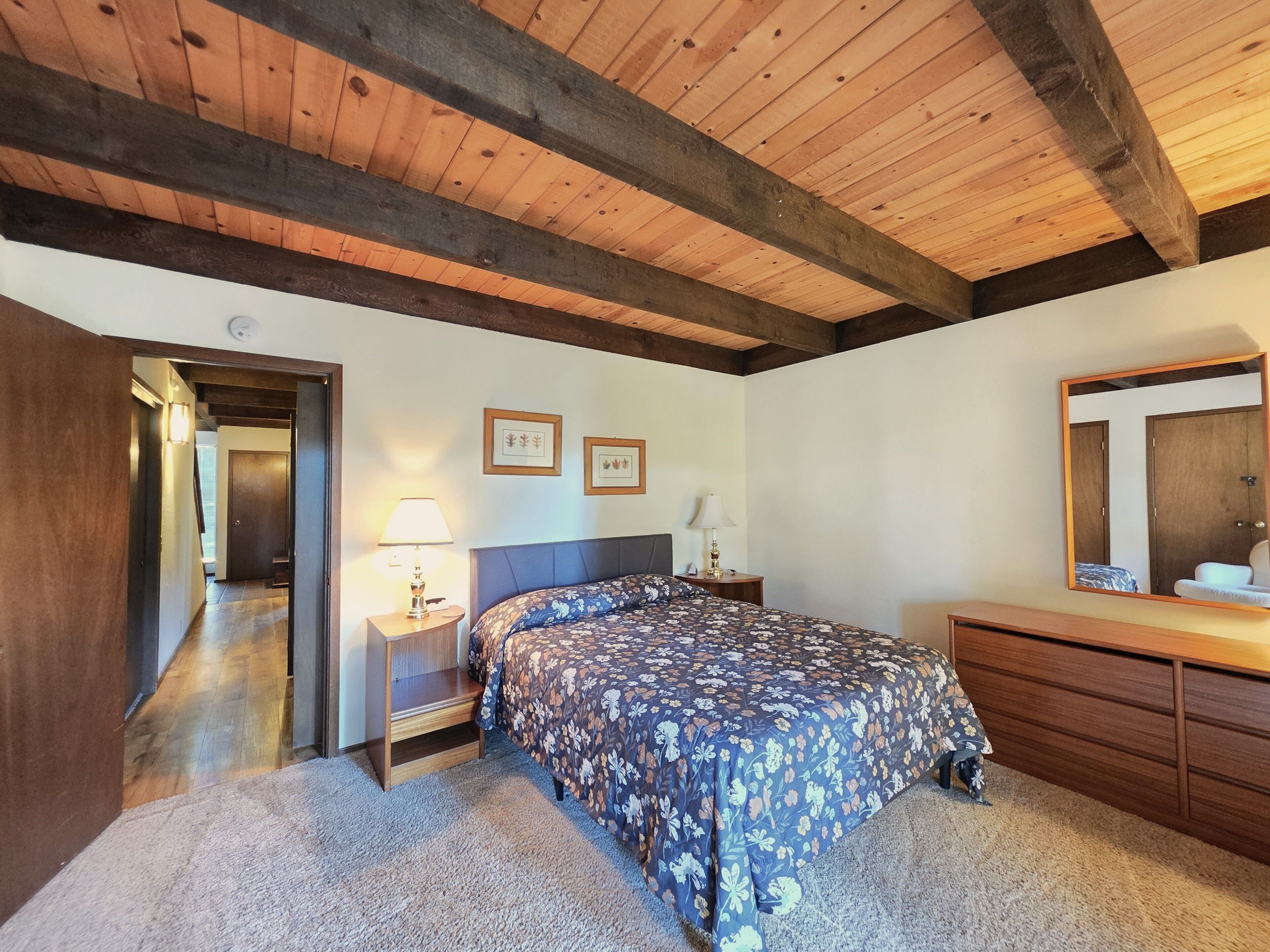 Une chambre avec un plafond en bois, un lit recouvert d’une literie à motifs floraux, une table de chevet avec une lampe, une commode et un miroir.