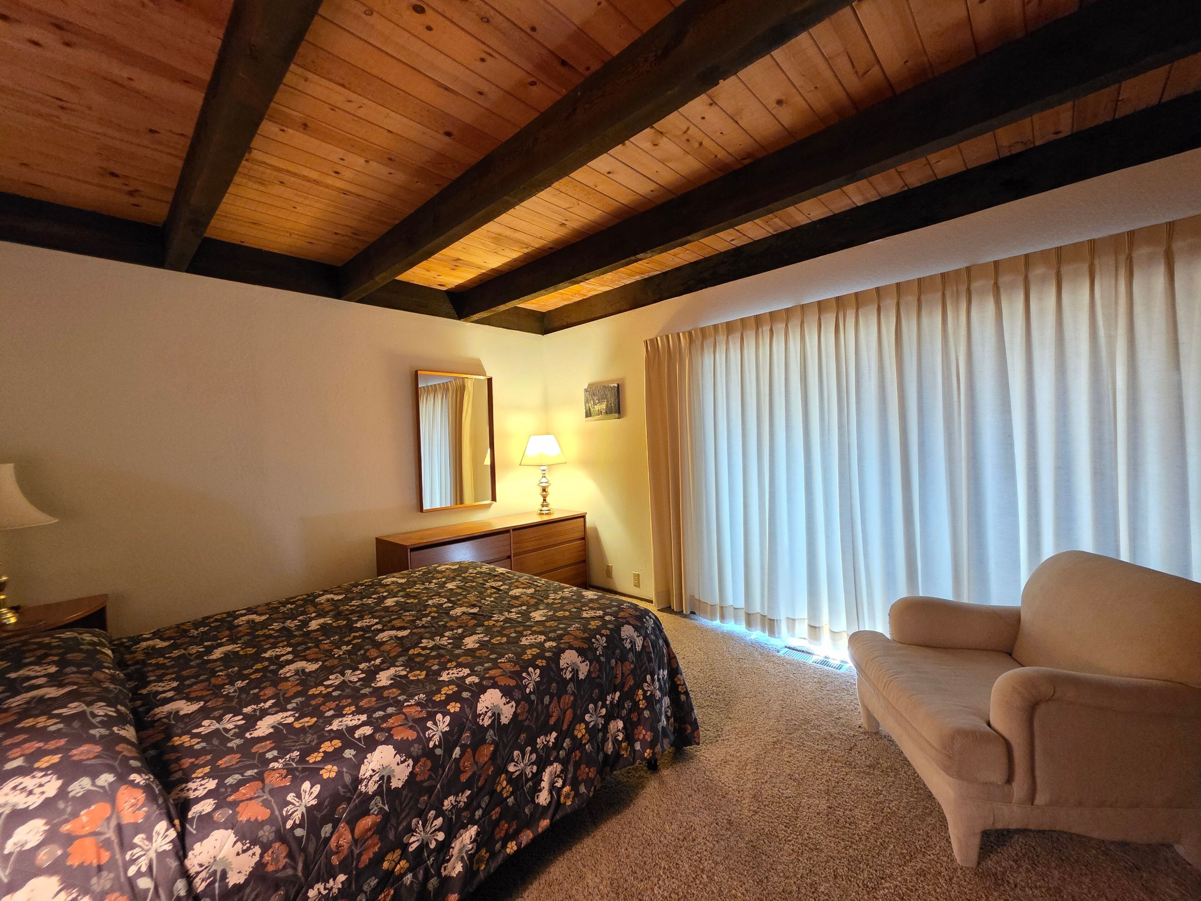 Un dormitorio con cama, una silla, una mesita de noche, una lámpara, un espejo y una ventana con cortinas.