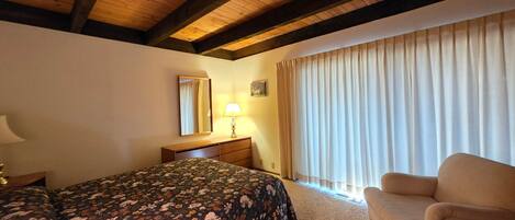 Un dormitorio con cama, una silla, una mesita de noche, una lámpara, un espejo y una ventana con cortinas.
