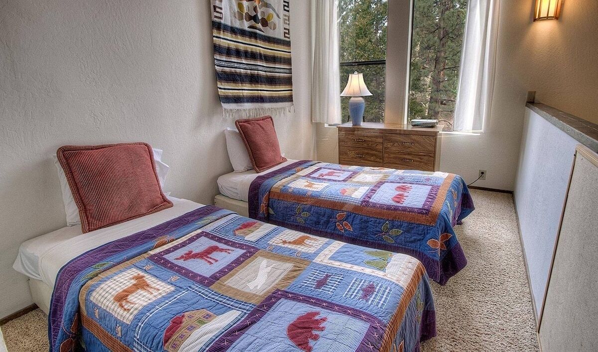 A bedroom with two beds, each with a quilt, a window with a view of trees, and a lamp on a nightstand.