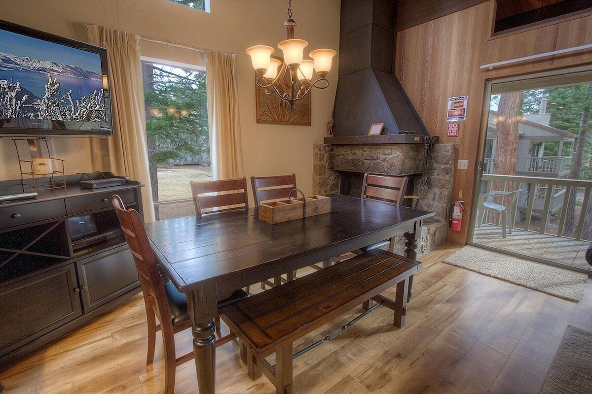 Un comedor con mesa de madera, sillas, un banco, televisor, candelabro y chimenea de piedra.