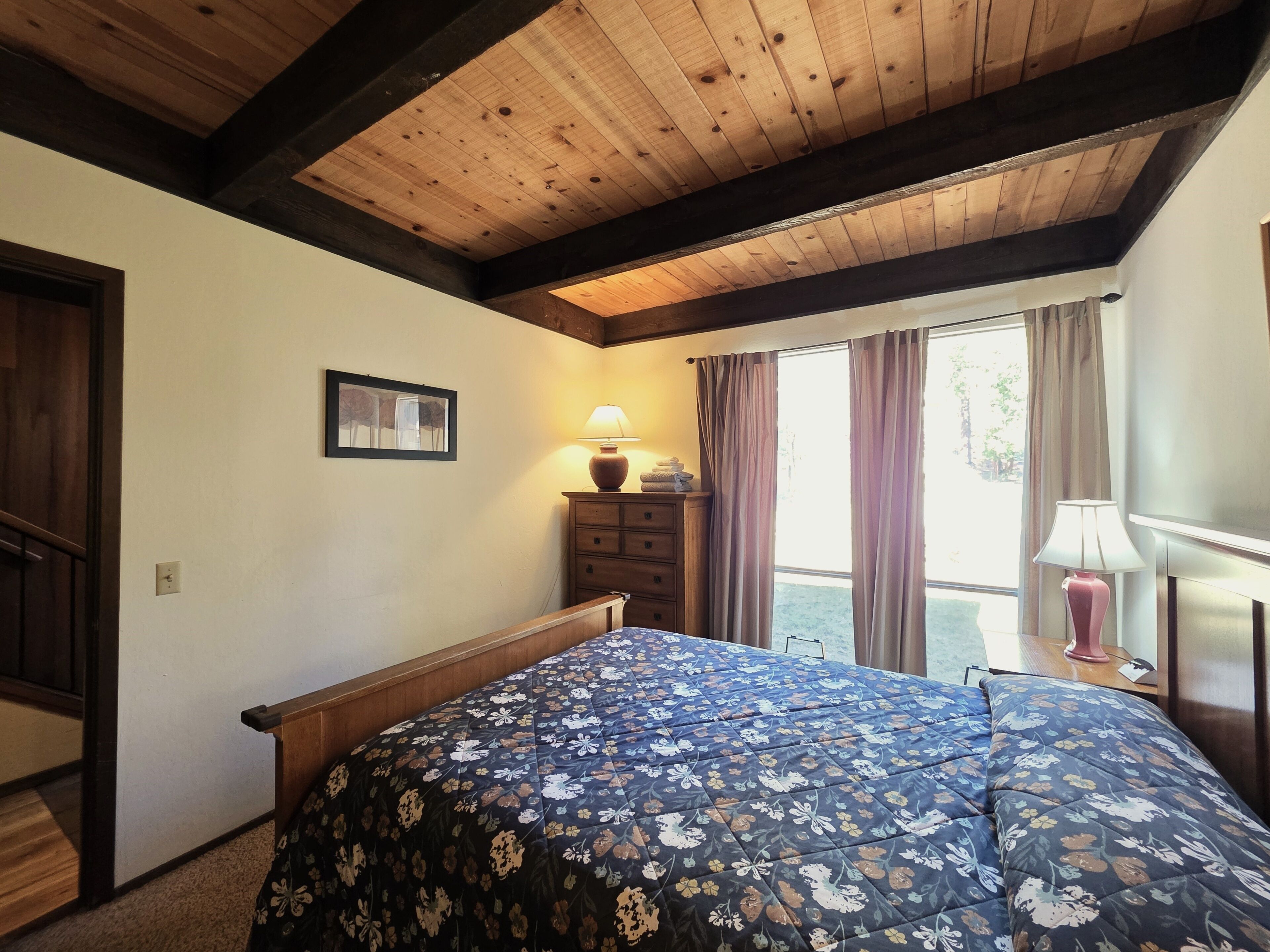 Un dormitorio con techo de madera, una cama con colcha floral, una mesita de noche con lámpara y una ventana con cortinas.