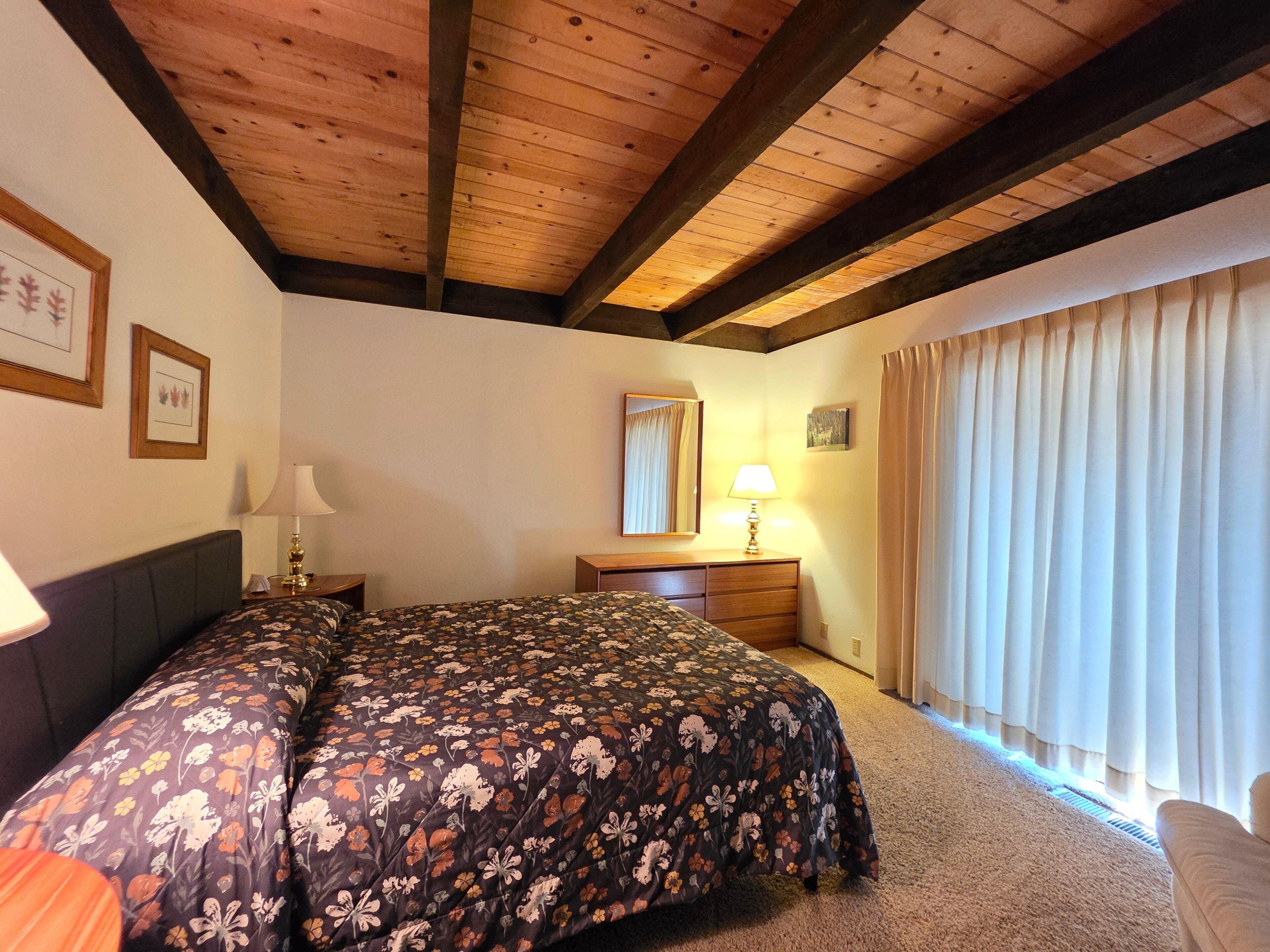 Ein Schlafzimmer mit Holzboden, einem Bett mit floralem Überwurf, einem Nachttisch mit Lampe, einer Kommode, einem Spiegel und einem Fenster mit Vorhängen.