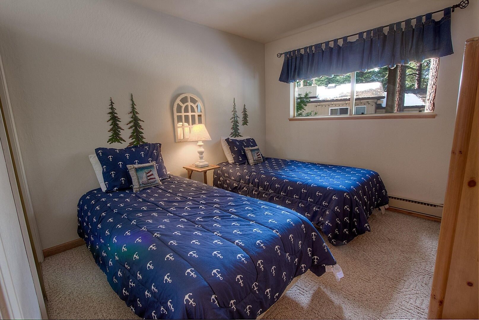 A bedroom with two beds, each with matching blue bedding and nautical-themed pillows, a small bedside table with a lamp, and a window with a blue valance.