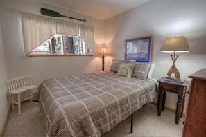 Un dormitorio con cama, ventana con cortinas, mesita de noche con lámpara, una silla y un cuadro en la pared.