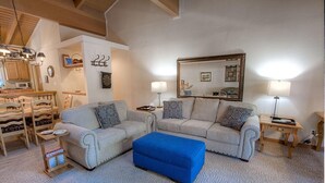 Una sala de estar con dos sofás, un puf azul, un comedor con sillas de madera y una cocina con microondas.