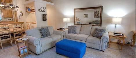 A living room with two sofas, a blue ottoman, a dining area with wooden chairs, and a kitchen with a microwave.