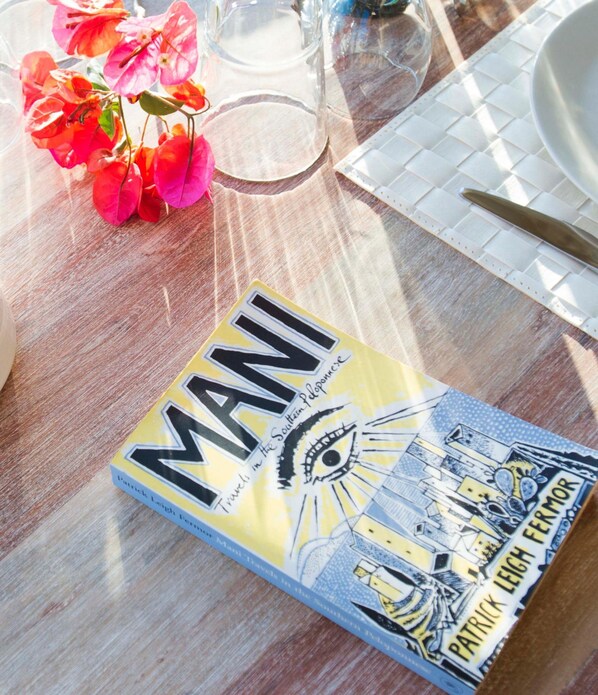 Um livro com o título 'MANI' sobre uma mesa de madeira com tampo de vidro transparente, rodeado por um jogo de toalha branco, um copo e uma tigela.