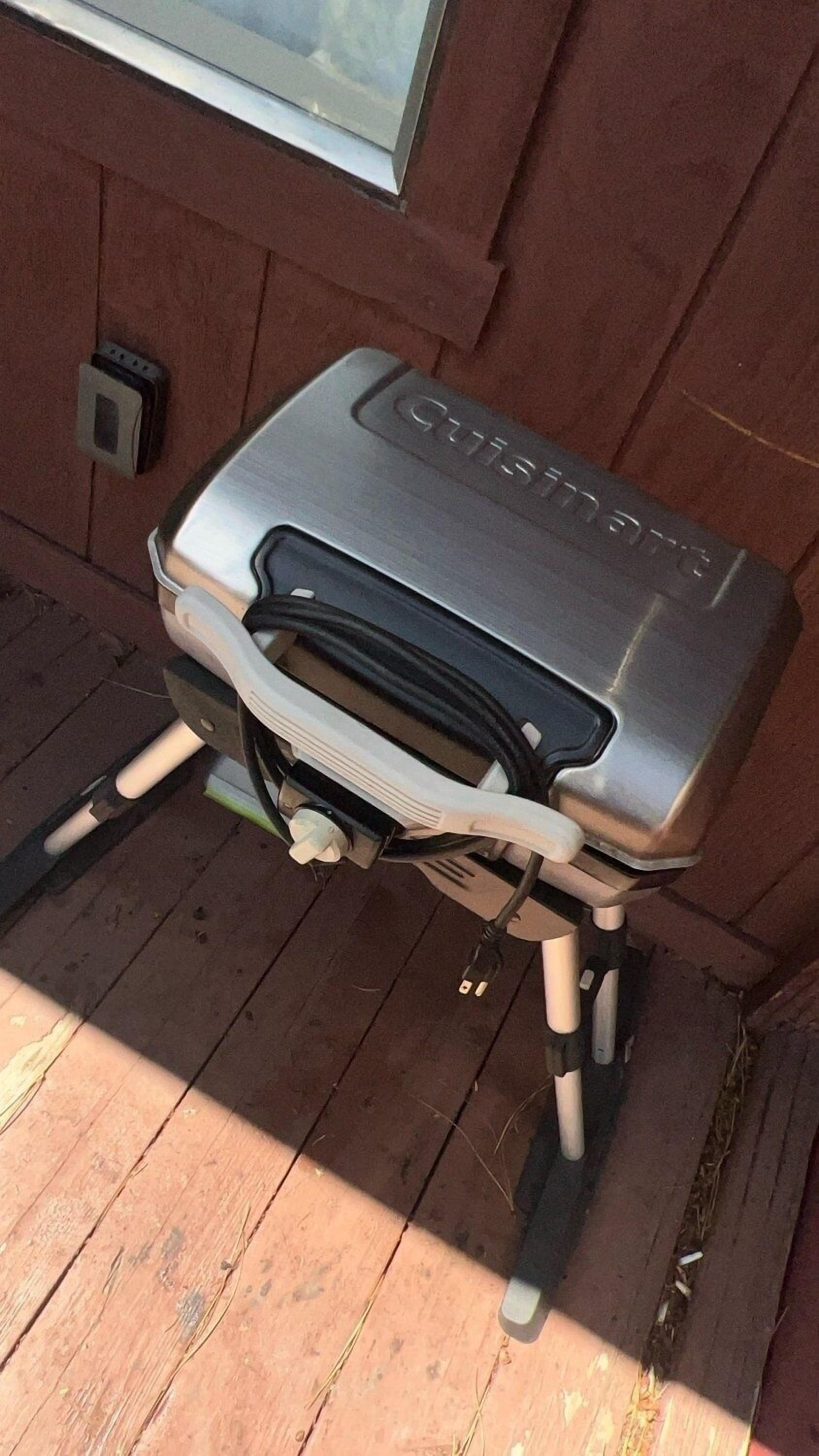 A portable grill with a handle and wheels, placed on a wooden deck against a brick wall.