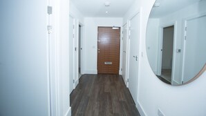A hallway with white walls, a wooden door at the end, and a round mirror on the right wall. - Highview 2-bed Apartment in London (London)