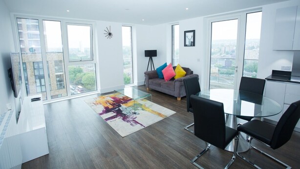 A modern living room with a glass dining table, grey sofa with colorful cushions, and a city view through large windows. - Highview 2-bed Apartment in London (London)