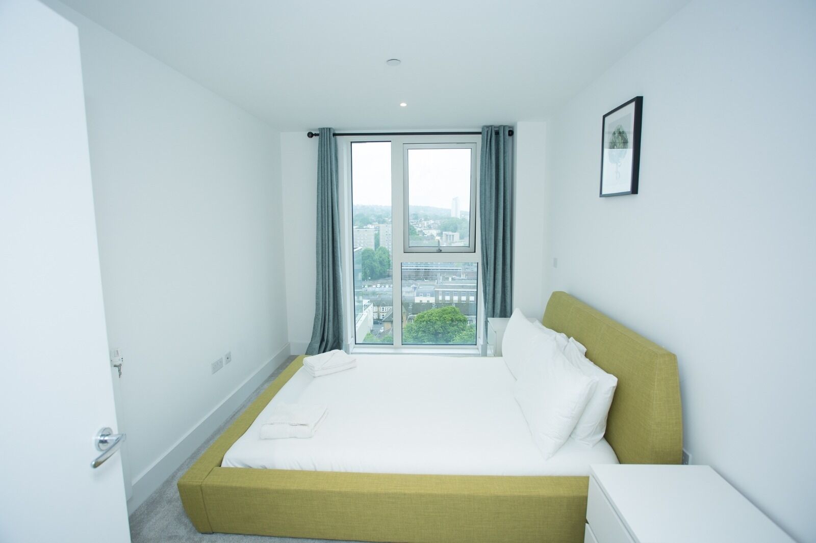 A hotel room with a large bed, a view of the city through a window, and a single framed picture on the wall.