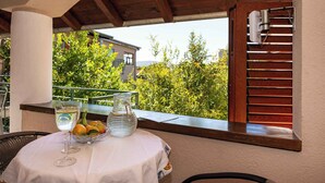 Een tafel met een glazen pitcher, een glas water en een schaal fruit op een balkon met houten leuningen en uitzicht op groen en gebouwen.