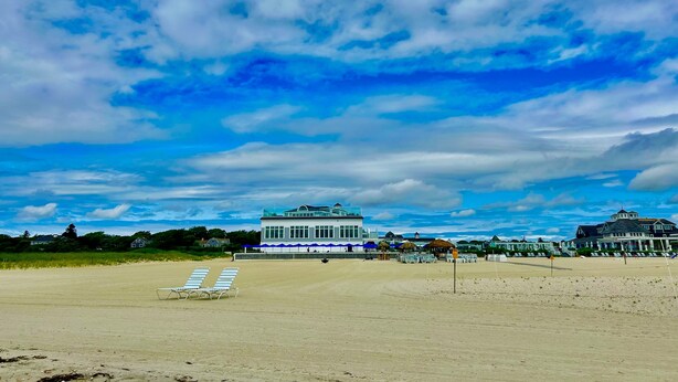 Una playa con un edificio grande al fondo, una zona de arena y un cielo despejado con nubes.
