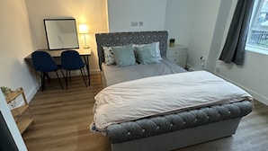 Ein Schlafzimmer mit einem Bett, einem Schreibtisch, zwei Stühlen, einem Spiegel und einem Fenster mit Vorhängen.