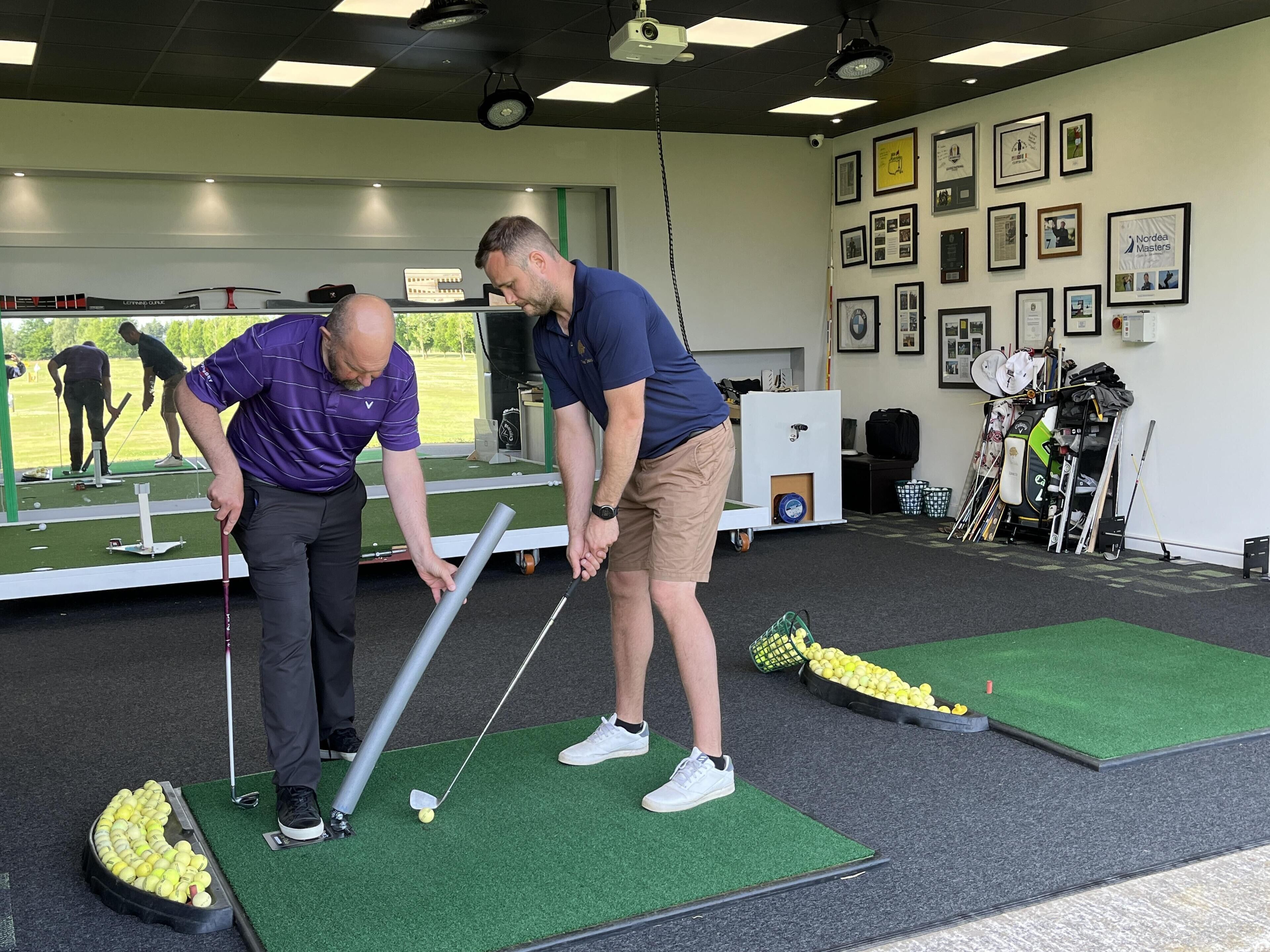 A golf training facility with multiple putting greens, a practice area, and a wall of framed photographs.