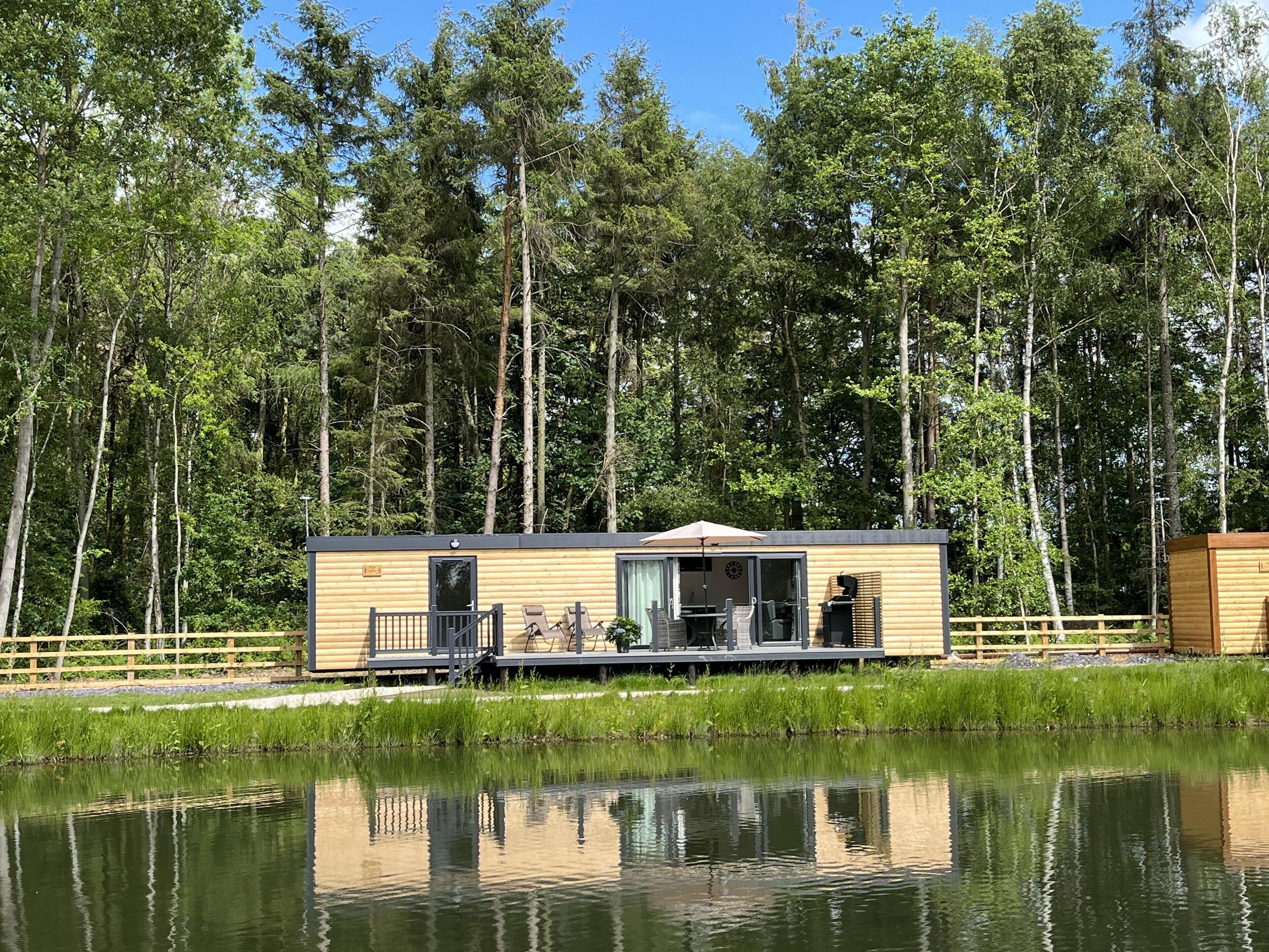 A cabin by a lake with a deck, a covered patio, and a view of the surrounding forest.