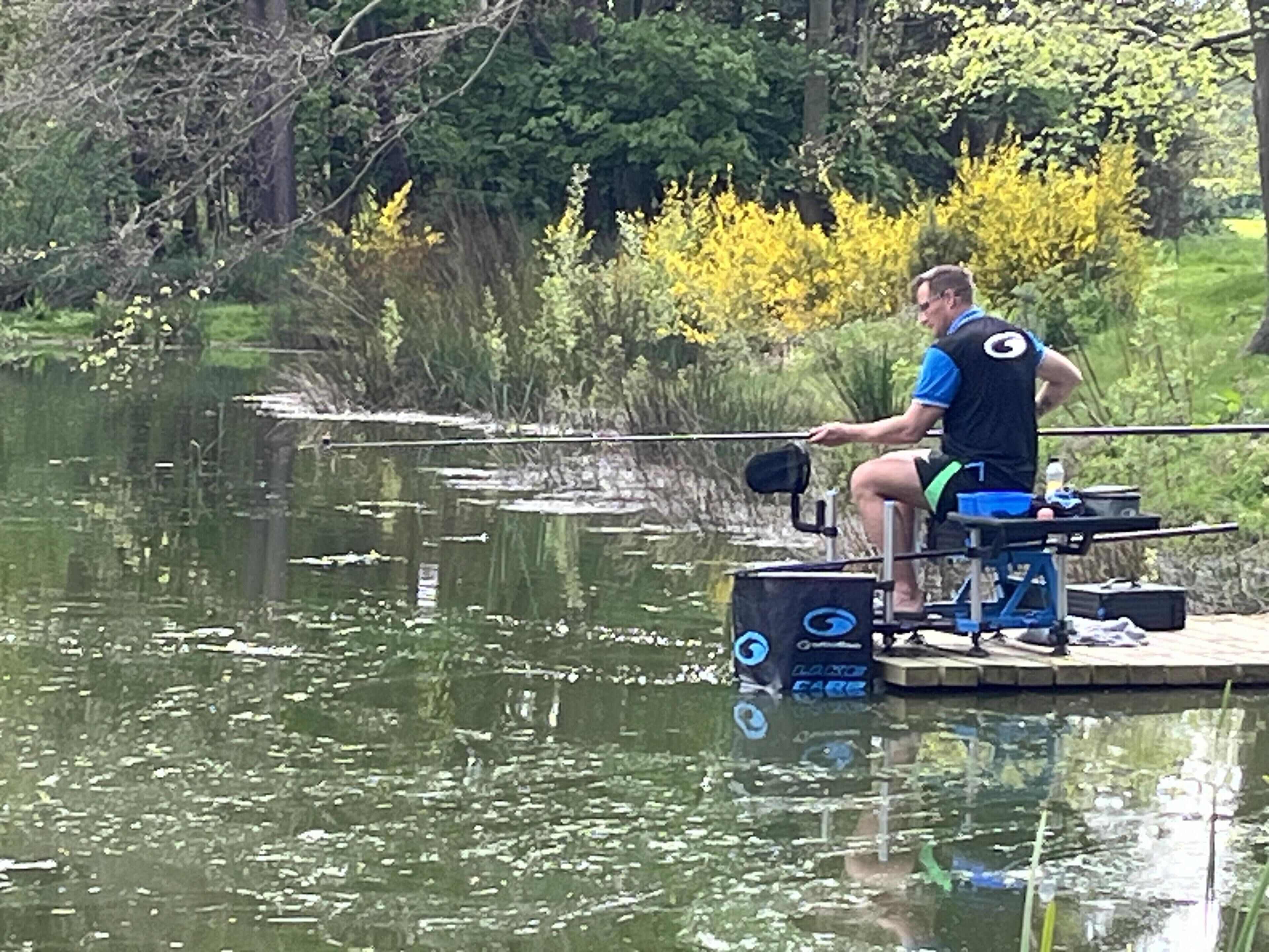 Une personne pêchant depuis une plateforme fixe, au bord d’une étendue d’eau calme, entourée de verdure et d’arbres.