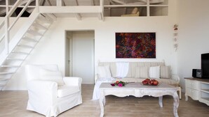 A spacious living room with a white sofa, a coffee table with flowers, a painting on the wall, and a staircase.