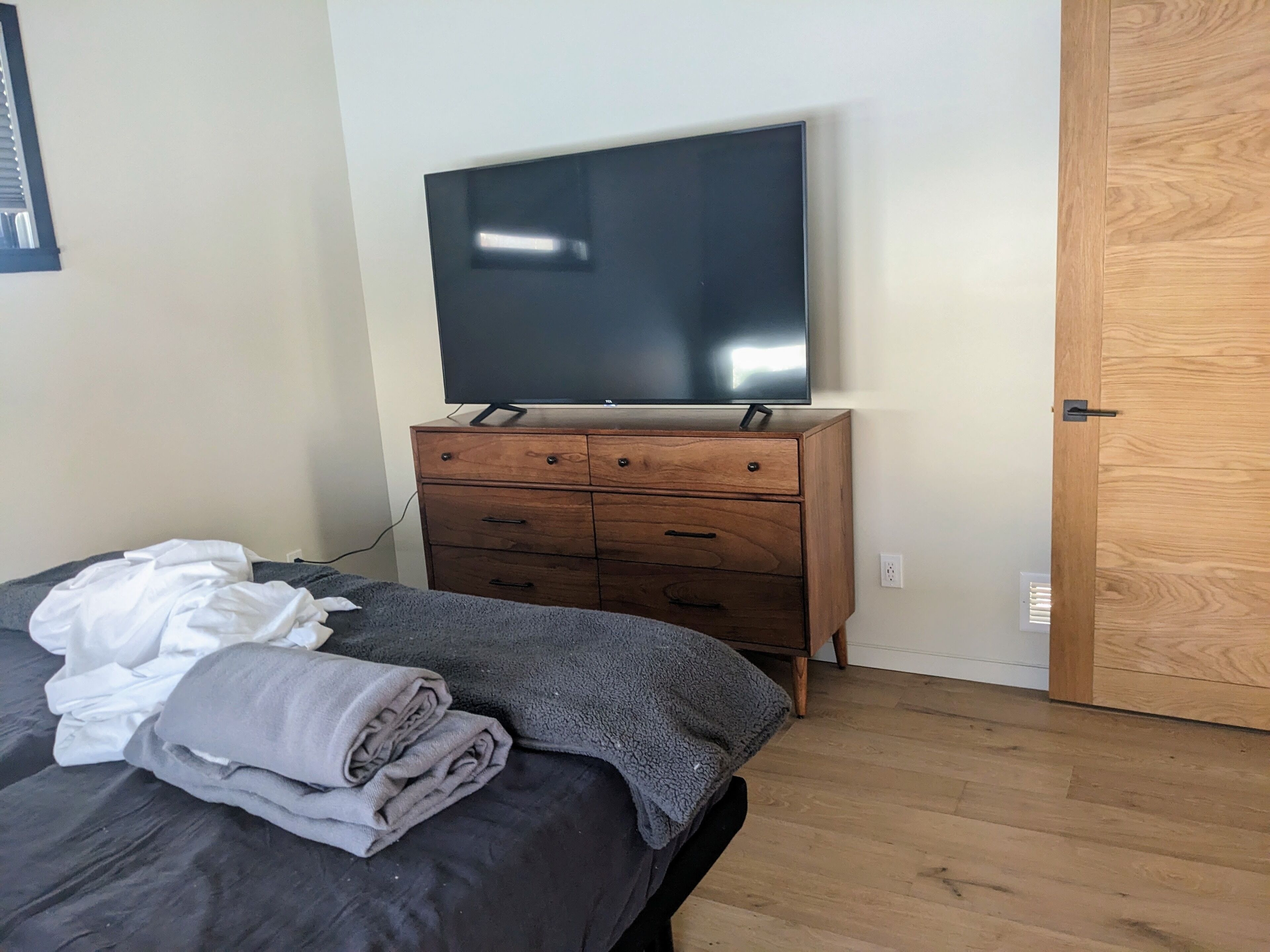 Une chambre à coucher avec un lit, une commode en bois surmontée d’un téléviseur à écran plat, une porte et une prise de courant murale.
