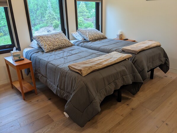 A double bed with grey bedding and a beige throw blanket, a bedside table with a remote control and tissues, and a window with a view of trees.