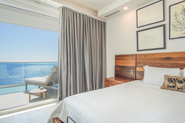 A bedroom with a large bed, a wooden headboard, a nightstand, and a view of the ocean.