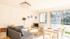 Een moderne woonkamer met een grijze bank, een eettafel met stoelen, een televisie en een groot raam met uitzicht op bomen.