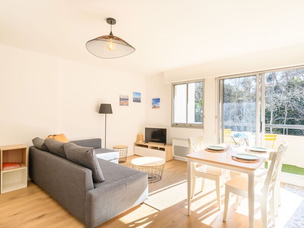 A modern living room with a grey sofa, a dining table with chairs, a TV, and a large window offering a view of trees.