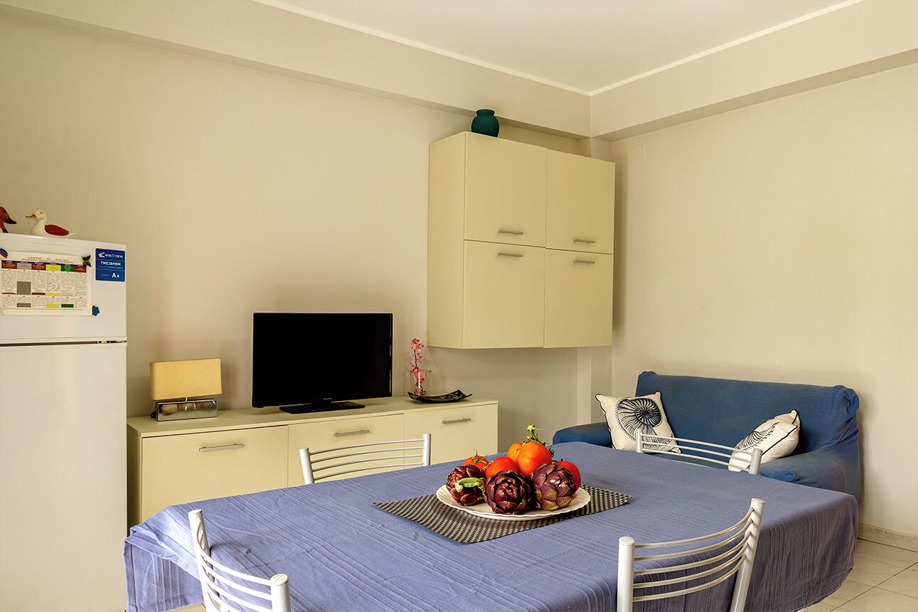 Une petite pièce, bien rangée, avec une table à manger dressée et ornée de légumes, un téléviseur posé sur un meuble et un canapé bleu.
