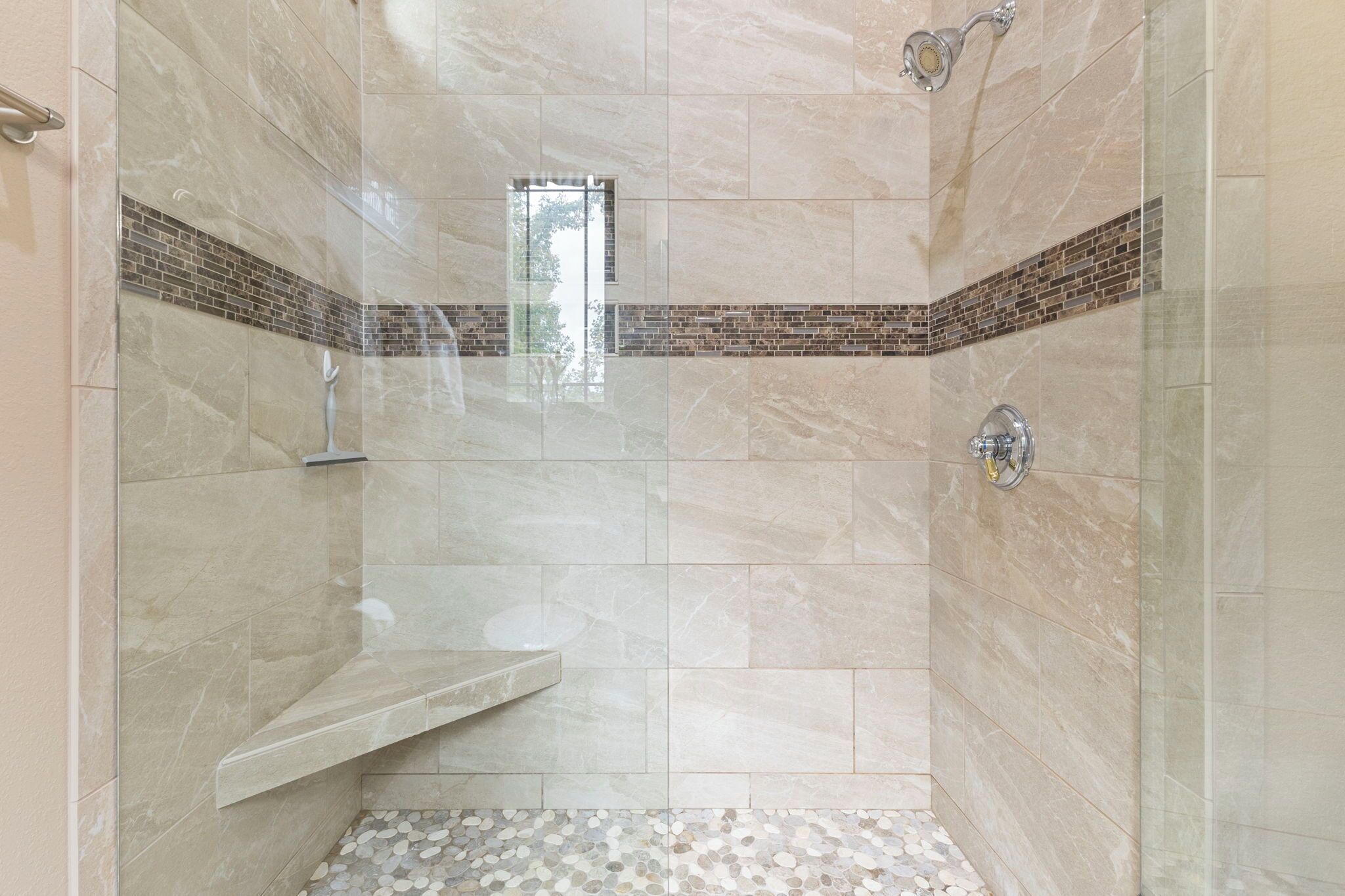 A modern bathroom with a glass shower enclosure, a small window, and a pebble-patterned floor.