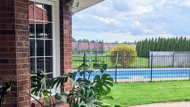 A brick building with a window and a potted plant in the foreground. In the background, there is a swimming pool surrounded by a fence and trees. - A poolside guest stay nested in a beautiful orchard along winery route (Niagara-on-the-Lake)