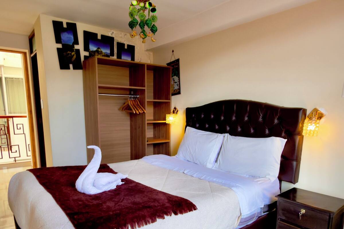 Een keurig opgemaakt bed met een handdoek in de vorm van een zwaan, een donker hoofdbord, een nachtkastje en een wandplank met decoratieve objecten.