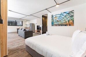 A modern hotel room with a large bed, a sofa, a TV, and a painting of a greenhouse.